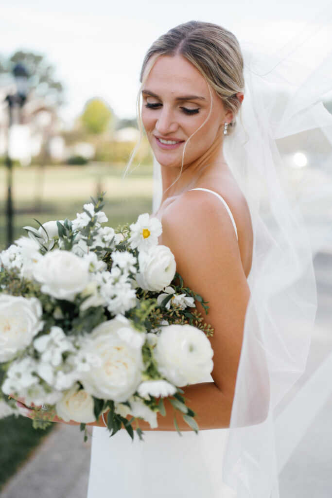 Bride smiling over her shoulder while holding a large bouquet of white flowers, wearing a veil and a sleek wedding dress outdoors on a sunny day. engagement party invitations