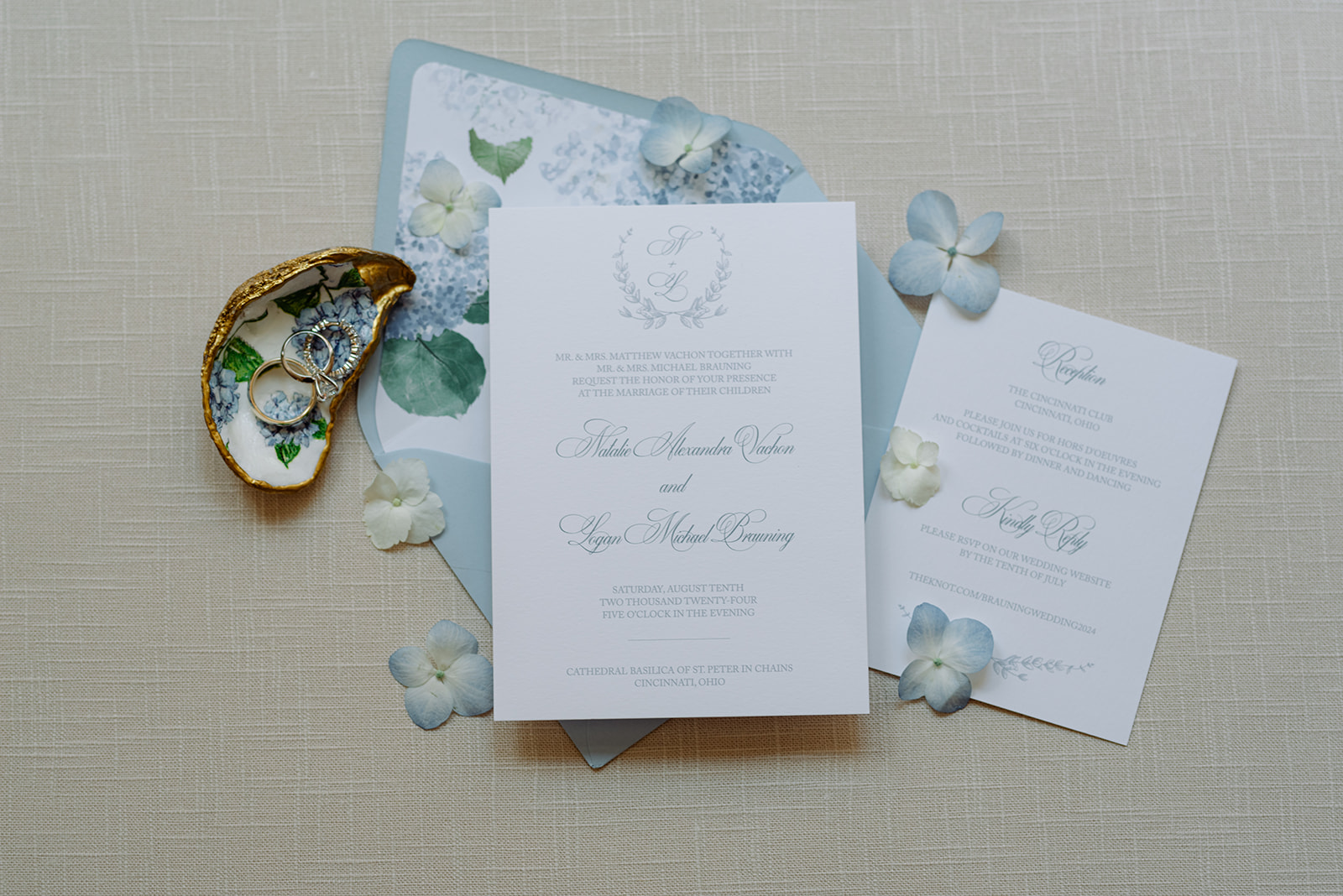 Flat lay of a classic white and blue wedding invitation suite with floral envelope liner, RSVP card, and delicate hydrangea petals, styled with gold-trimmed oyster shell holding wedding rings.