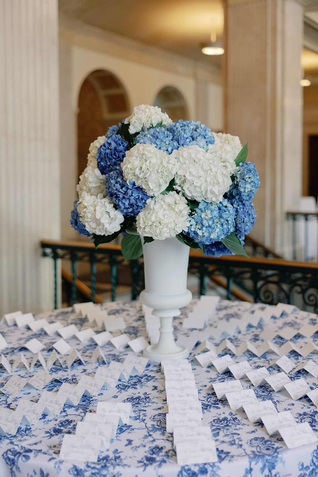 Escort card table featuring a lush blue and white hydrangea arrangement and toile-patterned linen. Rows of folded day-of stationery place cards add classic charm and refined organization to the space.