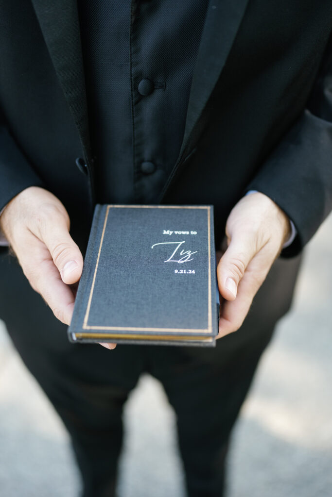 The groom holds a custom black vow book, adding a timeless, personal touch to his attire. 